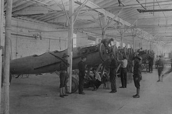 A line of airplanes is being assembled under the watchful eye of a gun-toting soldier as they are being completed to be sent to the European war; the factory is at College Point