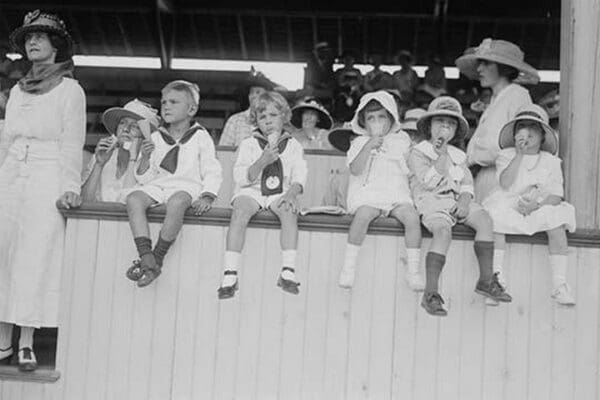 Children sit on wall in front of stands at the ballpark and eat ice cream cones. - Art Print