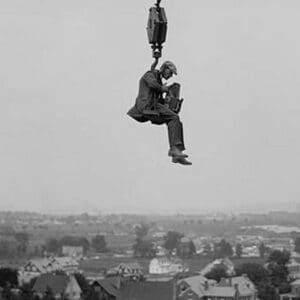 Still Photographer hangs suspended high above the ground on the hook of a large crane. - Art Print